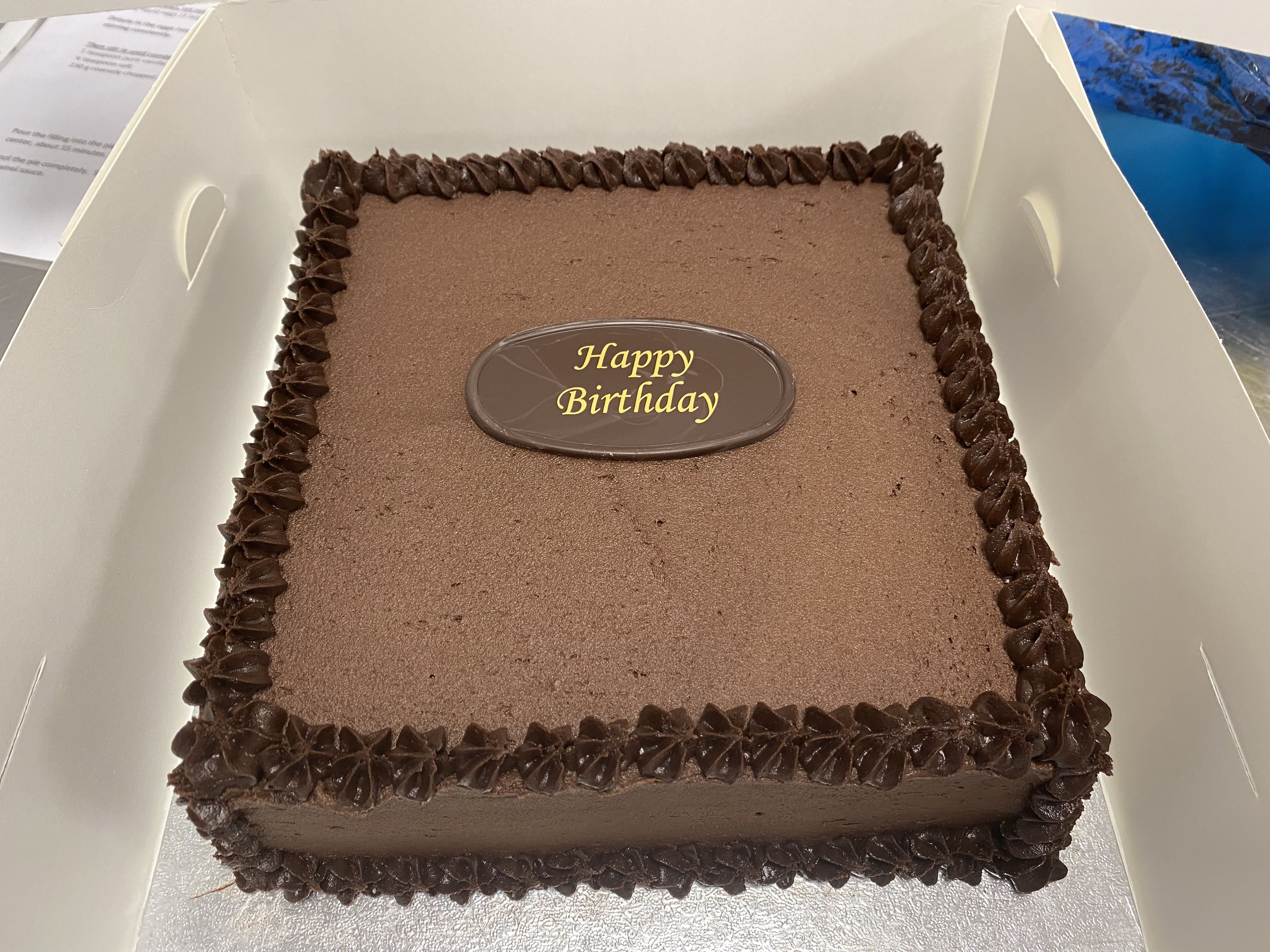 A decadent chocolate cake with velvety chocolate frosting edges is placed inside a white box. The cake boasts a smooth surface adorned with a chocolate plaque in the center that reads 'Happy Birthday' in vibrant yellow letters.
